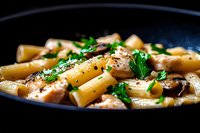 Creamy rigatoni pasta with mushrooms and fresh parsley in black pan, seasoned with black pepper and herbs, close up view of homemade Italian cuisine.