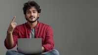 Young South Asian man in red jacket working on laptop while sitting cross-legged, gesturing with raised finger against gray background, expressing innovative idea.