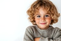 Young child with curly blonde hair and freckles wearing gray sweater smiling warmly at camera against white background, expressing genuine joy and innocence.