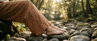 Young woman legs in brown pants walking barefoot on smooth river stones in forest setting with natural sunlight filtering through trees creating peaceful outdoor scene.