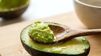 Fresh ripe avocado half with wooden spoon containing mashed guacamole on natural kitchen counter for healthy cooking and meal preparation.