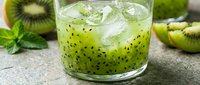 Refreshing kiwi cocktail with ice in a glass, surrounded by fresh kiwi slices and mint leaves on a stone surface.