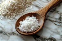 Coarse sea salt crystals in wooden spoon on marble surface, natural light illuminates white mineral grains, raw cooking ingredient for food preparation.