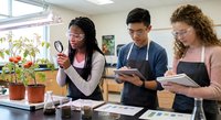 Three diverse high school students conducting plant biology experiment in modern science laboratory classroom with magnifying glass and notebooks.