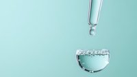 Water drop falling from dropper into small glass container with clear liquid on mint green background. Minimalist composition for cosmetic and skincare advertising.