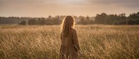 Young woman with long hair walking through golden wheat field at sunset, peaceful countryside landscape with warm lighting for wellness and nature concepts.