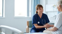 Female nurse in blue scrubs with stethoscope providing compassionate care to elderly patient in hospital room, healthcare professional consultation.