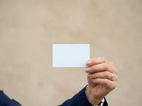 Mature man in business attire holding blank white card against neutral beige wall background, suitable for corporate branding, business card mockups, and professional marketing materials.