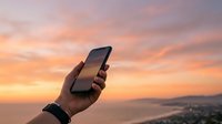 Hand holding smartphone against sunset sky with coastal landscape below, capturing moment of digital connection and travel photography.