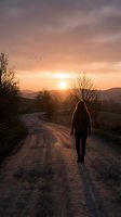 Woman with long reddish-brown hair, wearing dark clothing, walking alone on rural dirt road at golden hour sunset with mountains and bare trees in background.