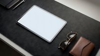 Tablet with blank white screen on dark felt desk mat beside leather wallet and glasses, modern workspace mockup for app or web design.