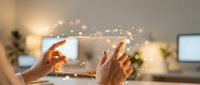 Woman's hands holding tablet device with glowing digital particles and light effects floating above screen in modern office workspace environment.