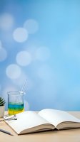 Colorful layered drink in glass with straw next to open book on wooden desk against blue bokeh background for summer reading and relaxation concepts.