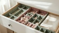 Organized jewelry drawer with velvet compartments holding rings, earrings, scrunchies and a watch in soft sage green and blush pink tones.