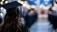 Young female graduate in black cap and gown with long brown wavy hair viewed from behind against blurred bokeh lights background at graduation ceremony.