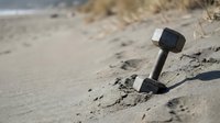 Single dumbbell weight lying on sandy beach near water, fitness equipment abandoned on shore for workout, exercise and training concept.
