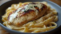 Grilled chicken breast with creamy alfredo sauce and fettuccine pasta, garnished with cracked black pepper and fresh herbs served on gray plate.