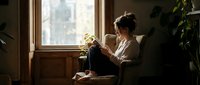 Young woman sitting by window reading book in cozy home interior with natural light and houseplants creating peaceful morning atmosphere for relaxation.