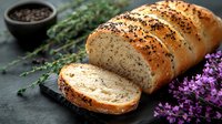Fresh baked artisan bread loaf with black sesame seeds, sliced on dark stone board, garnished with purple flowers and herbs, rustic food styling.