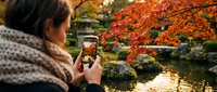 Woman with dark hair photographed from behind wearing cozy knit sweater, holding smartphone to capture autumn foliage. Vibrant orange and red maple trees frame serene water landscape. Fall season travel and nature photography.