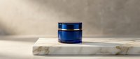 Blue glass cosmetic jar on marble platform against textured beige wall background for skincare product photography and beauty brand marketing campaigns.