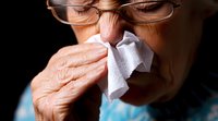 Senior woman with glasses blowing nose into tissue paper against dark background, showing cold and flu symptoms in dramatic lighting. Healthcare concept.