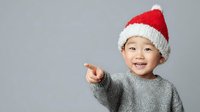 Cheerful child wearing Santa hat and gray sweater pointing with finger against neutral background. Holiday season portrait.