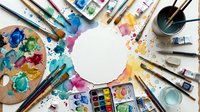 Overhead view of artist workspace with blank white circle surrounded by watercolor paints, brushes, palettes, and art supplies on colorful splattered surface for creative projects.