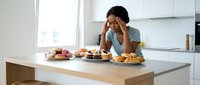 Young African American woman experiencing stress while sitting at kitchen counter surrounded by unhealthy junk food and sweets during emotional eating episode.