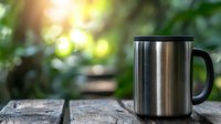 Stainless steel travel mug on rustic wooden surface against blurred natural green background with sunlight bokeh, morning coffee or tea concept for outdoor lifestyle.