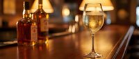 Glass of white wine on wooden bar counter with whiskey bottles in background creating warm ambient lighting for restaurant and hospitality industry.