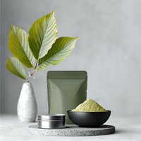 Fresh green powder in black bowl with metallic tin and pouch on stone platform, decorative leaves in white vase against gray background for wellness products.