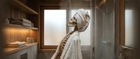 Woman with wrapped head towel standing in modern bathroom with wooden shelving, warm lighting, and glass shower enclosure, suitable for spa, wellness, and home design content.