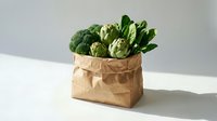 Fresh organic vegetables broccoli and artichokes in eco-friendly brown paper bag on white table. Sustainable grocery shopping and healthy eating concept.