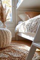 Cozy bedroom corner with chunky knit pouf, textured rug, and white bedding, decorated with dried pampas grass. Natural sunlight creates warm atmosphere in minimalist space.