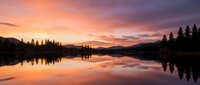 Serene lake at sunrise with golden and pink sky reflecting on calm water, surrounded by evergreen forest and distant mountains, peaceful nature landscape.
