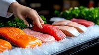 Fresh raw salmon and tuna sashimi pieces on crushed ice with hand placing fish, close-up view of premium seafood preparation in restaurant kitchen.