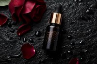 Luxury skincare serum bottle with rose gold dropper cap surrounded by red rose petals on dark textured stone surface for premium beauty and cosmetics branding.