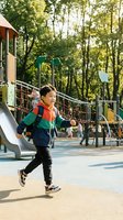 Happy Asian boy running at playground wearing colorful jacket during sunny day with climbing equipment and trees in background for active childhood concepts.