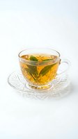 Clear glass teacup with saucer containing golden herbal tea and fresh green leaves on white background for wellness and beverage concepts.
