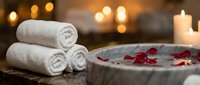 Luxury spa bath setup with rolled white towels, rose petals in water, and glowing candles creating romantic wellness atmosphere for relaxation and self-care.