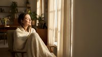 Young woman relaxing in cozy knit blanket by sunny window at home enjoying peaceful morning light and warmth in comfortable living space.