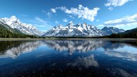 Majestic snow-capped mountain peaks reflecting in crystal clear alpine lake waters under bright blue sky with white clouds, surrounded by evergreen forest landscape.
