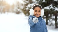 Happy African American girl in blue winter coat and white earmuffs pointing at camera in snowy park with warm sunlight and evergreen trees in background.