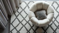 Cozy round cream-colored armchair with plush cushioning on geometric patterned gray rug, modern minimalist living room interior with soft natural lighting.