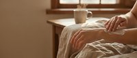 Person relaxing in cozy bedroom with warm morning sunlight streaming through window, hands resting on soft bedding creating peaceful atmosphere.