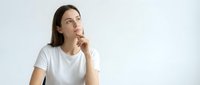 Young thoughtful woman in white t-shirt looking up with finger on chin in contemplative pose against light background for business concepts.
