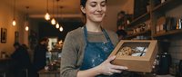 Young woman with short dark hair and fair complexion, wearing a blue apron over casual clothing, holding an open kraft paper box in a warm-lit retail or café environment. Suitable for small business, e-commerce, packaging, and customer service concepts.
