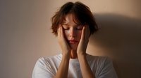 Young Asian woman in white t-shirt holding head in hands showing signs of stress, anxiety, depression or headache against neutral wall background in natural lighting.