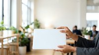Businesswoman holding blank white card in modern office with colleagues and plants in background for corporate messaging and branding.